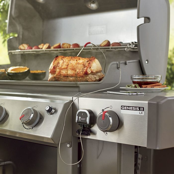 A grill is cooking a large piece of meat and vegetables while displaying temperature settings, set outdoors with smoke rising in a sunny environment.