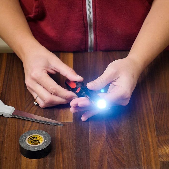 A person holds a flashlight, illuminating their hands while seated at a wooden table. Scissors and tape are nearby, suggesting a crafting or repair task.