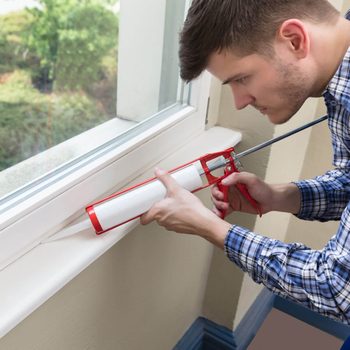 A man uses a caulking gun to apply sealant around a window, focusing intently in a well-lit indoor space with greenery visible outside.