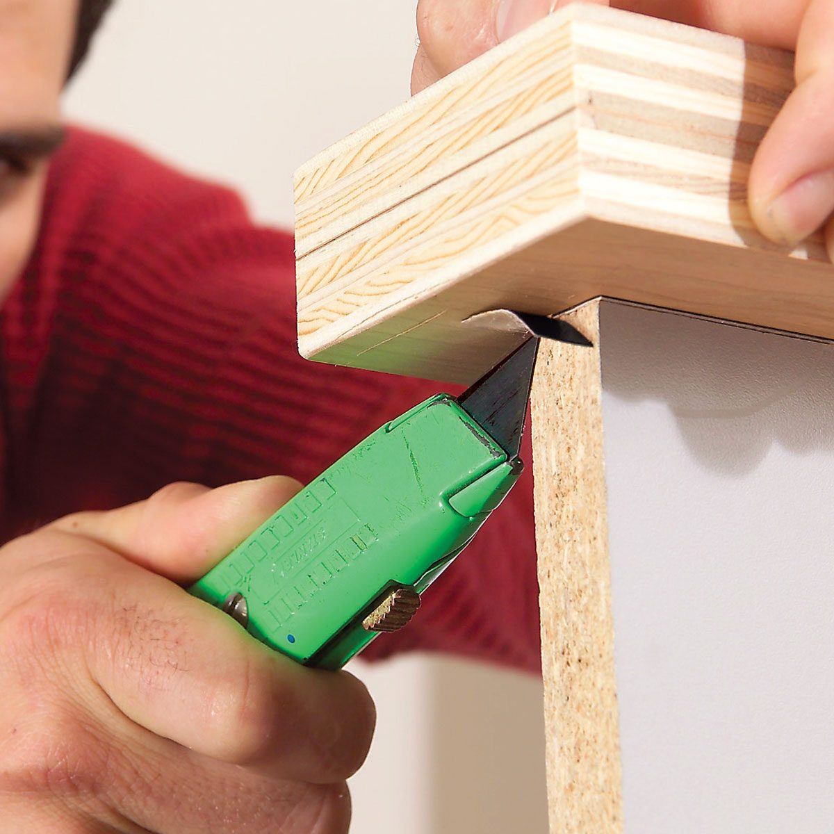 A hand holds a green utility knife, cutting the edge of wooden material, while a patterned, red fabric background suggests a casual indoor setting.