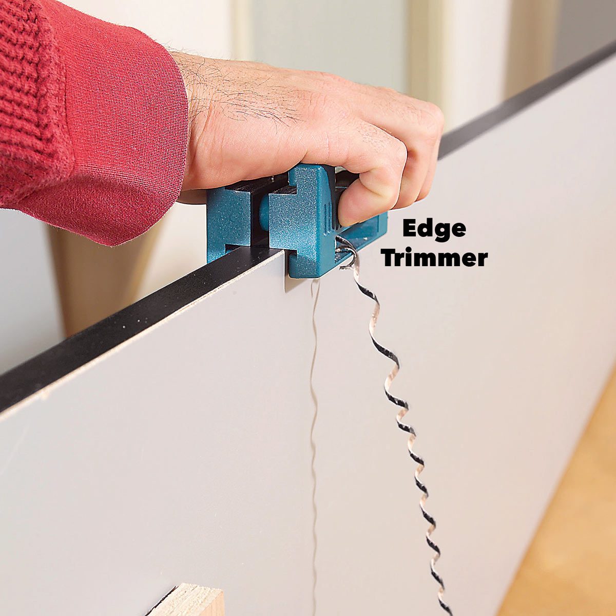A blue edge trimmer is held against a board's edge, while its spiral blade trims the surface. The background is lightly blurred, suggesting a workshop setting.