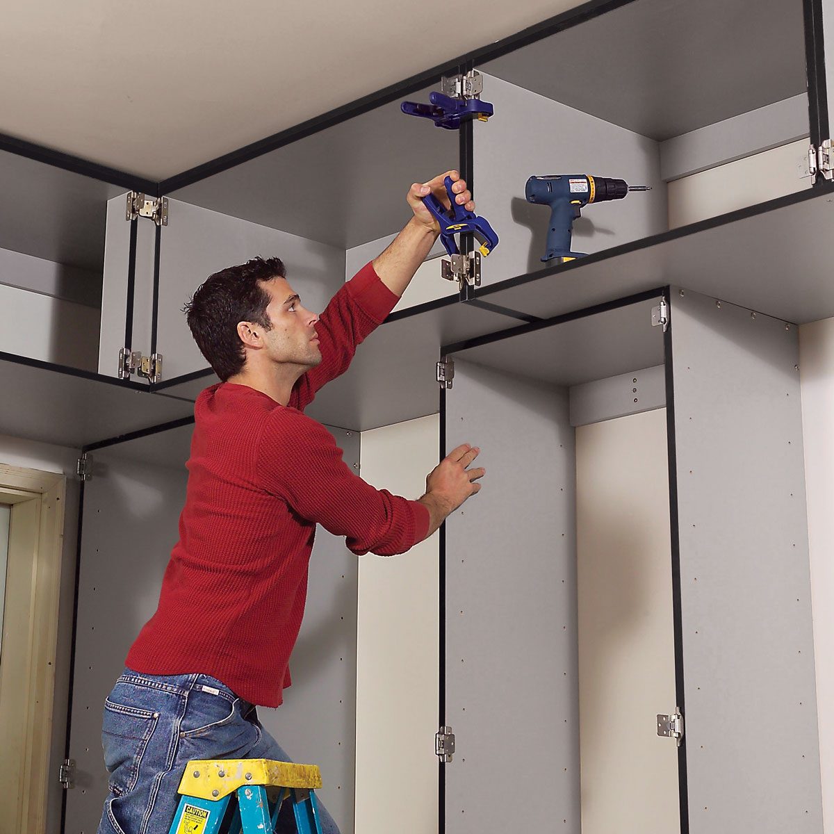 A man on a ladder is securing cabinets using a clamp and a drill, surrounded by unfinished cabinetry in a home.