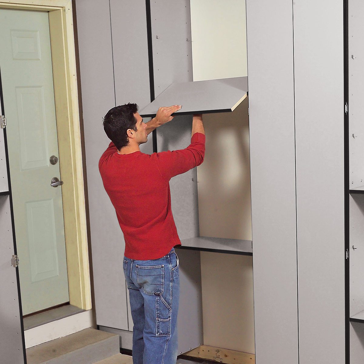 A man lifts a shelf to install it into a gray cabinet against a wall, next to a door in a home setting.