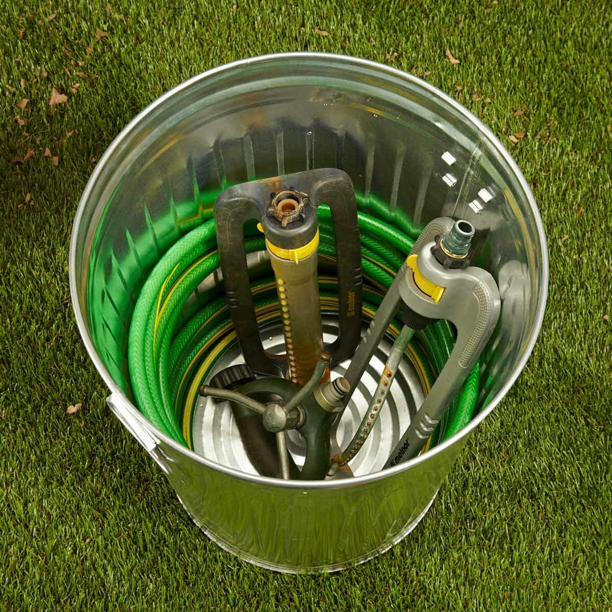 A metal bucket contains coiled green hoses and gardening tools, resting on a grassy surface under natural light.