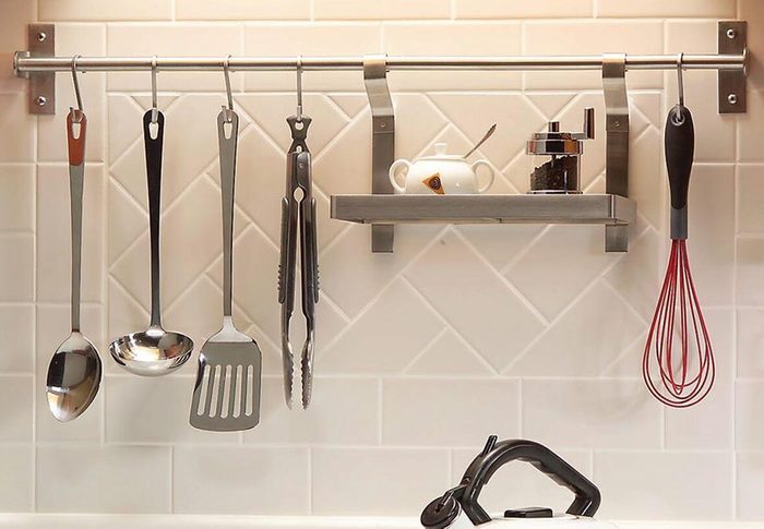 A set of cooking utensils hangs on a metal rack above a countertop, with a white kettle sitting nearby on a tiled kitchen surface.