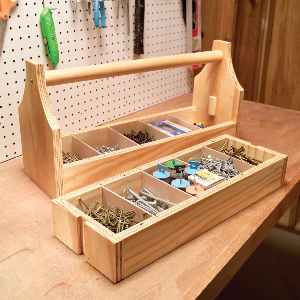 A wooden toolbox organized with various compartments holds screws, nails, and fasteners, resting on a workbench against a pegboard background.