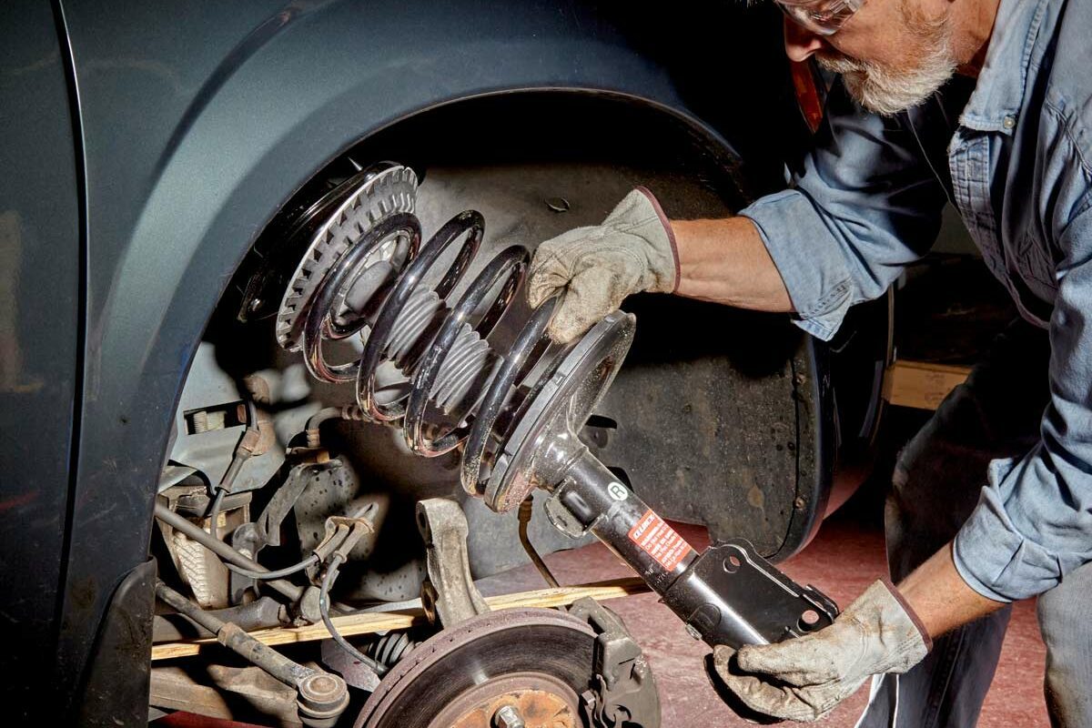 A man, wearing gloves and safety glasses, is removing a suspension coil from a vehicle in a well-lit garage, exposing the brake and suspension components.