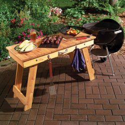 A wooden table displays grilled meats and corn, with a drink nearby, while a charcoal grill sits beside it in a garden setting.