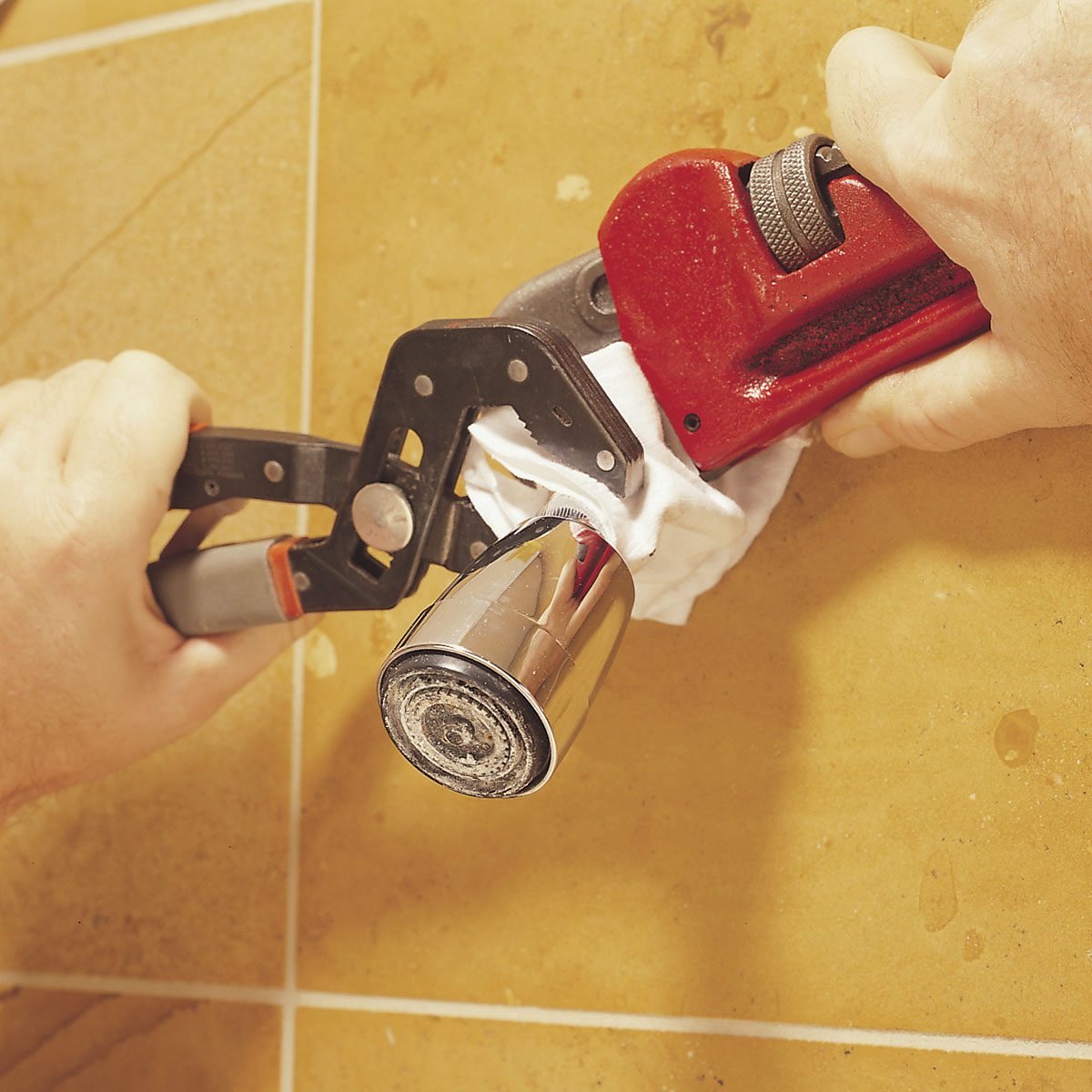 A hand uses a wrench to grip a silver faucet in a bathroom setting, with water-splattered tiles and a cloth positioned around the faucet.