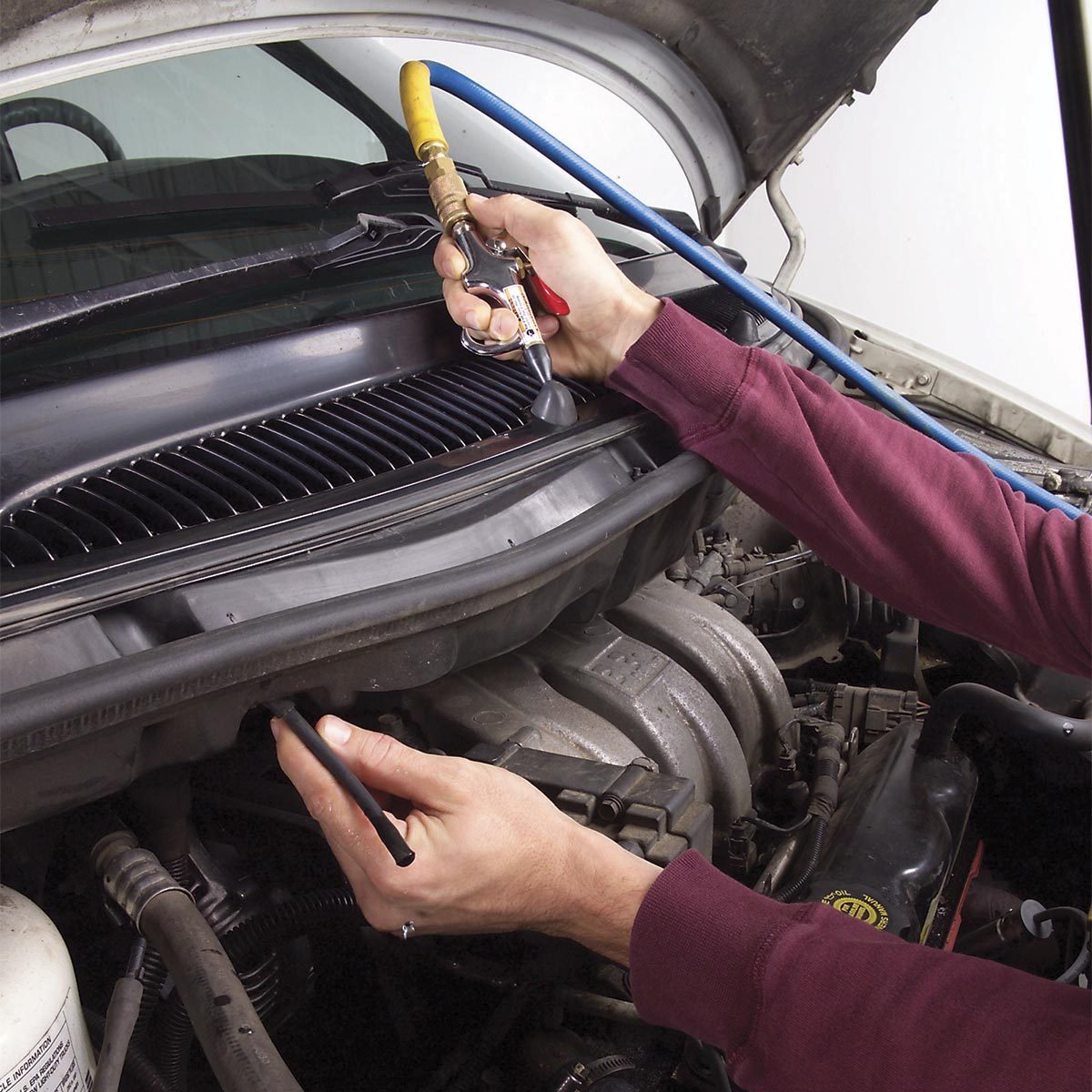 A hand holds an air nozzle, cleaning an engine compartment. A wrench is also in the other hand, surrounded by various car parts in a garage setting.