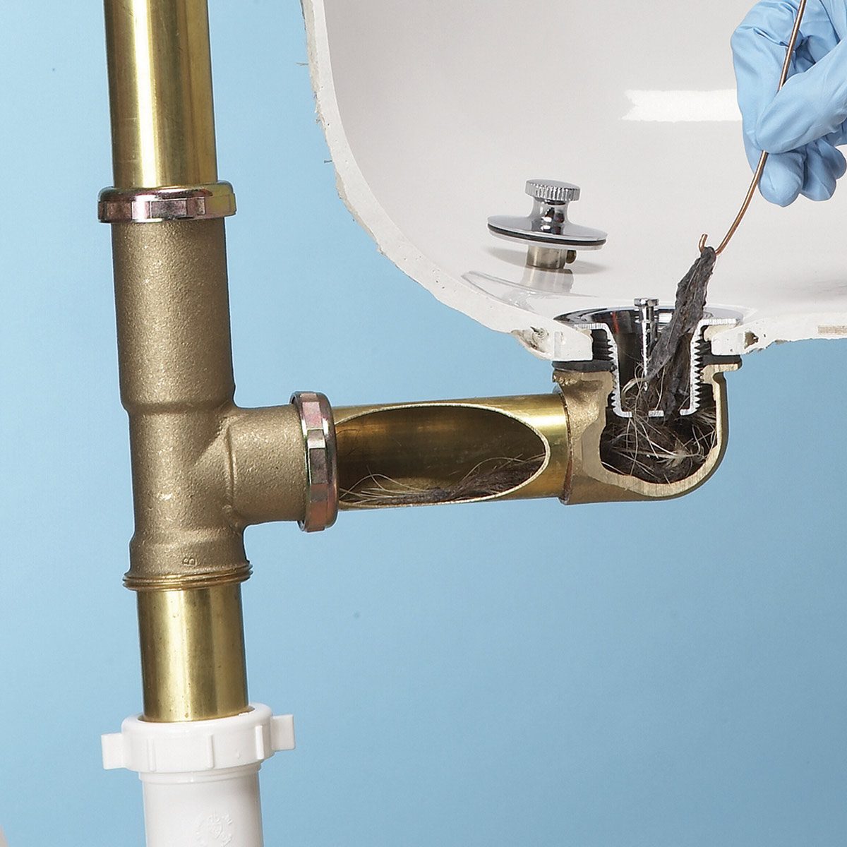 A person wearing a blue glove removes hair and debris from a plumbing pipe connected to a sink, set against a light blue background.