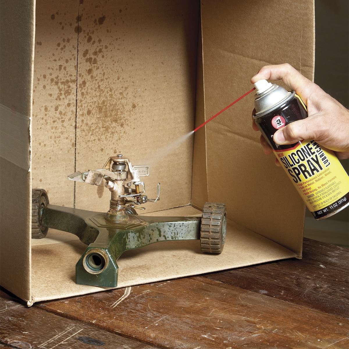 A hand sprays silicone lubricant onto a metal sprinkler in a cardboard box, with a wooden surface beneath, showcasing a controlled workspace for maintenance.