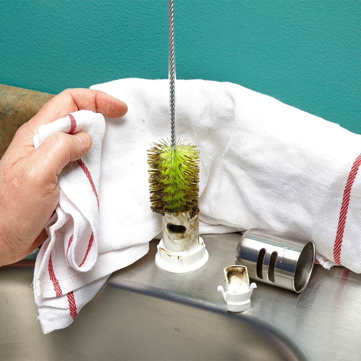 A hand holds a towel while removing a cleaning brush from a white sink attachment, surrounded by metal and plastic tools on a shiny surface.