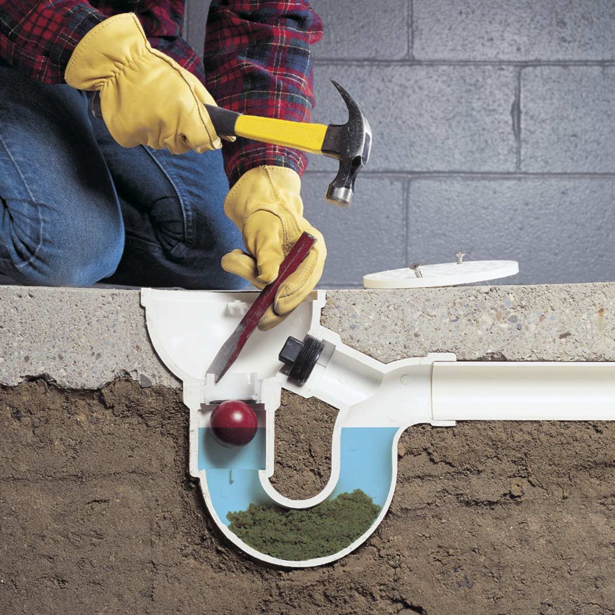A person in gloves uses a hammer on a plumbing component, revealing a cross-section with a ball and soil, surrounded by concrete and a gray wall.