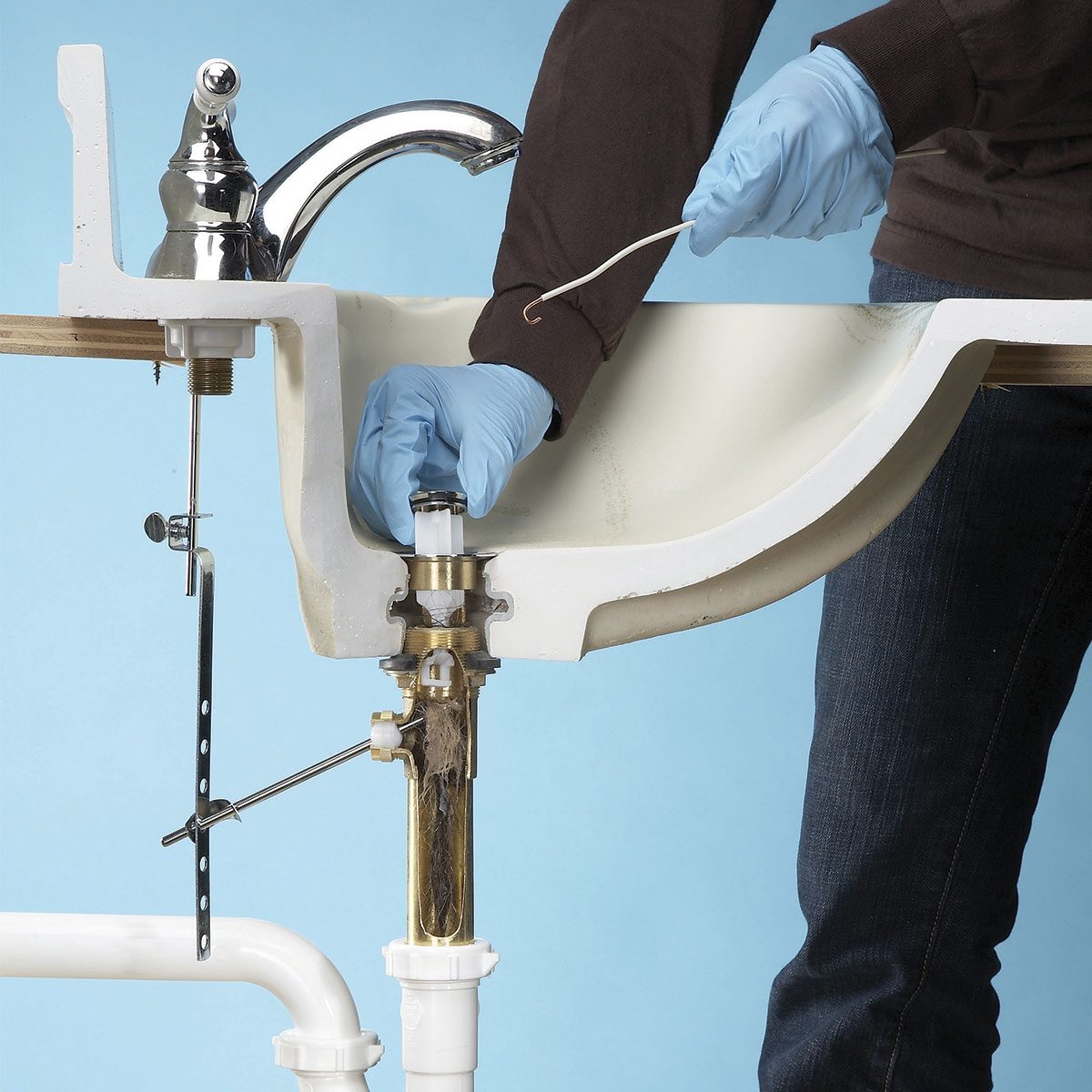 A person wearing gloves is installing a bathroom sink's drain assembly against a blue background, utilizing tools to secure the plumbing components.