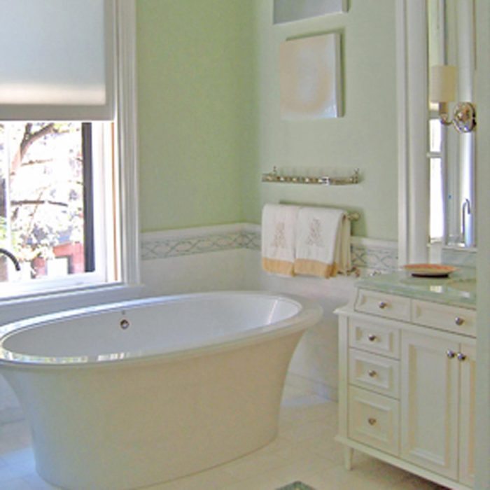 A white bathtub rests in a light green bathroom, beside a vanity with drawers and a sink, while towels hang above a decorative border.