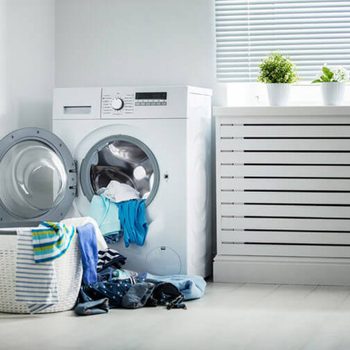A washing machine with an open door displays mixed laundry spilling out. Nearby, a laundry basket is filled with neatly folded clothes in a well-lit, modern room.