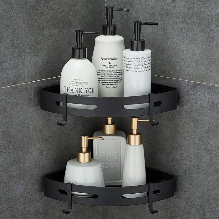 Two black corner shelves hold five white pump bottles in a bathroom setting, labeled with various names and decorative text against a gray tiled wall.