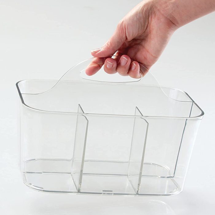 A clear plastic container is being lifted by a hand, designed with a handle and internal dividers, set against a light background.