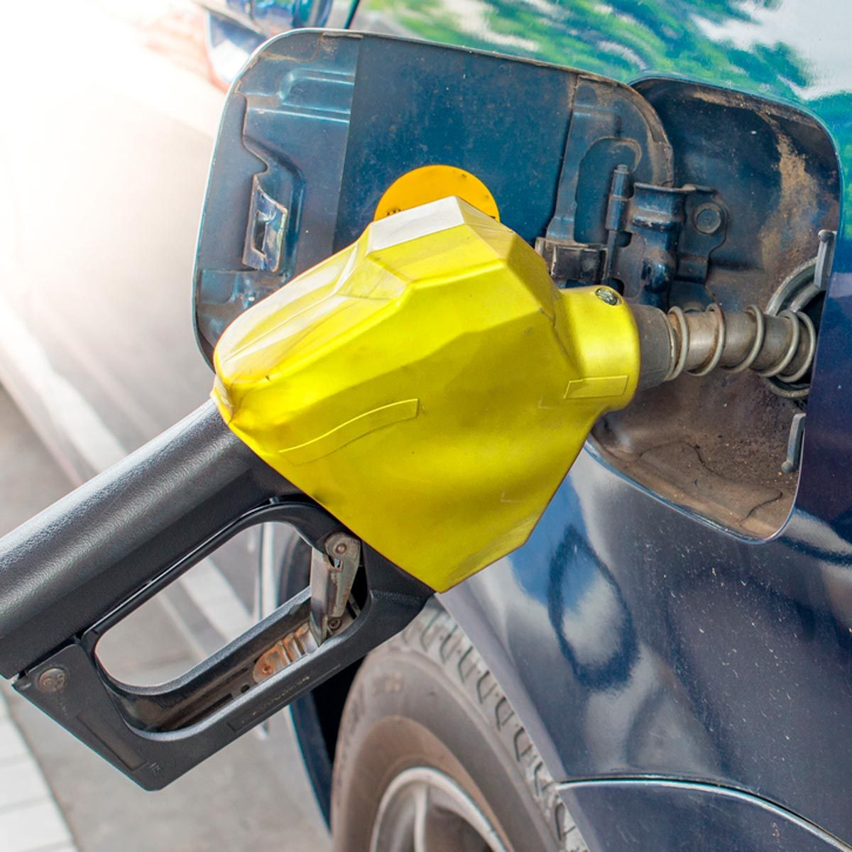 A yellow fuel nozzle is inserted into a car's gas tank, dispensing fuel at a gas station, with a blurred background of pavement and another vehicle.