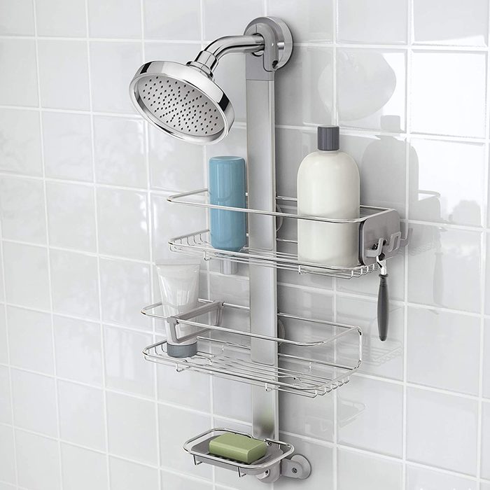 A chrome showerhead is mounted on a wall alongside a multi-tiered silver storage rack holding various toiletries in a tiled bathroom environment.