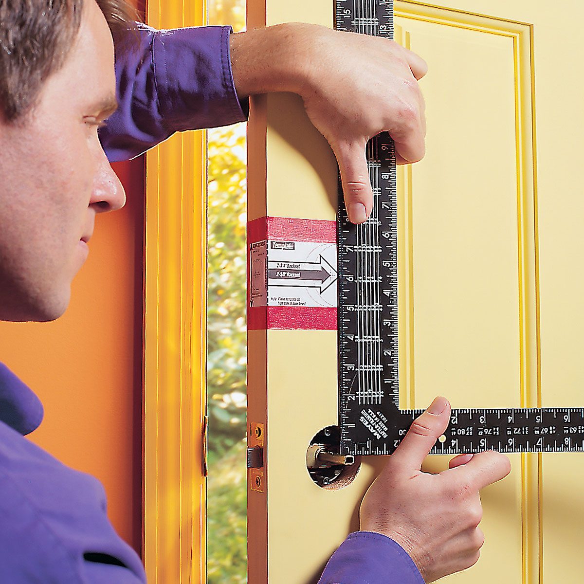 A person measures a yellow door frame using a framing square and tape measure, surrounded by an orange wall and a window with a garden view.