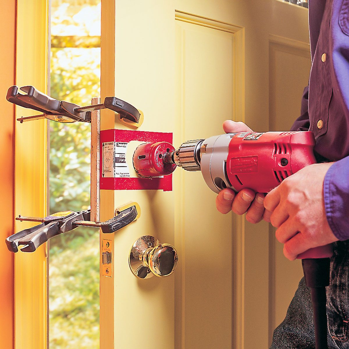 A person is using a power drill to install a door lock on a yellow door, secured with clamps, in a bright indoor setting.
