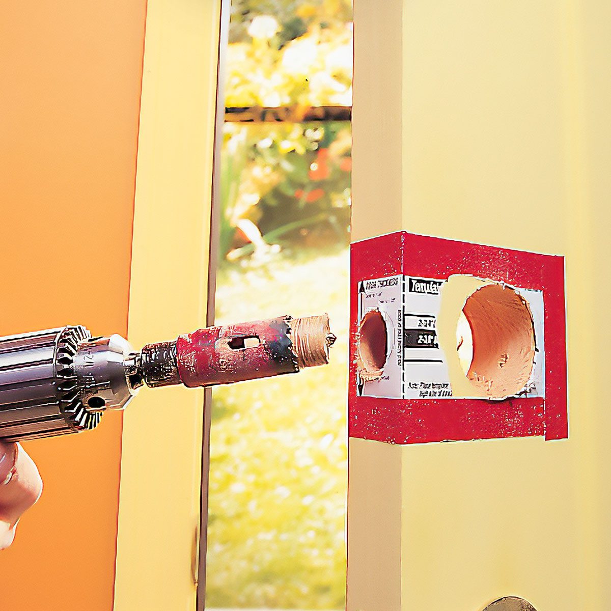A drill bit is boring a hole into a door frame, with a partially finished cut surrounded by a red template, set against a sunny outdoor background.