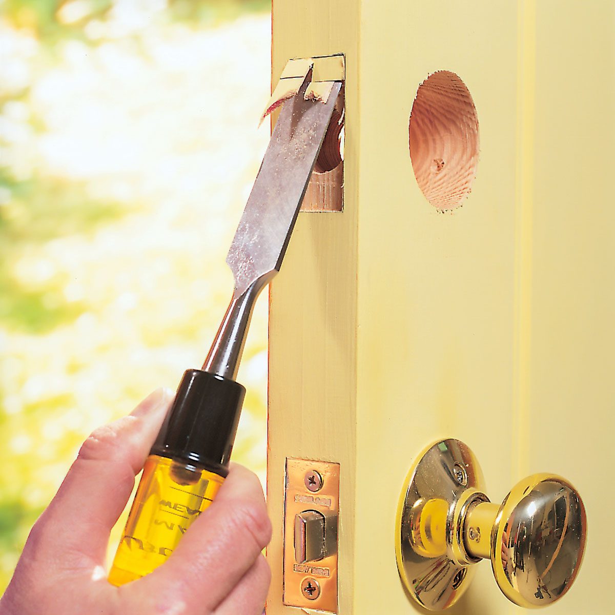 A chisel is being used to carve a door edge, with a hand gripping the tool. A polished doorknob and a warm, bright background are visible.