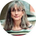 A woman with long, gray hair and glasses stands confidently, wearing a striped sweater, against a blurred urban background, suggesting a relaxed, outdoor setting.