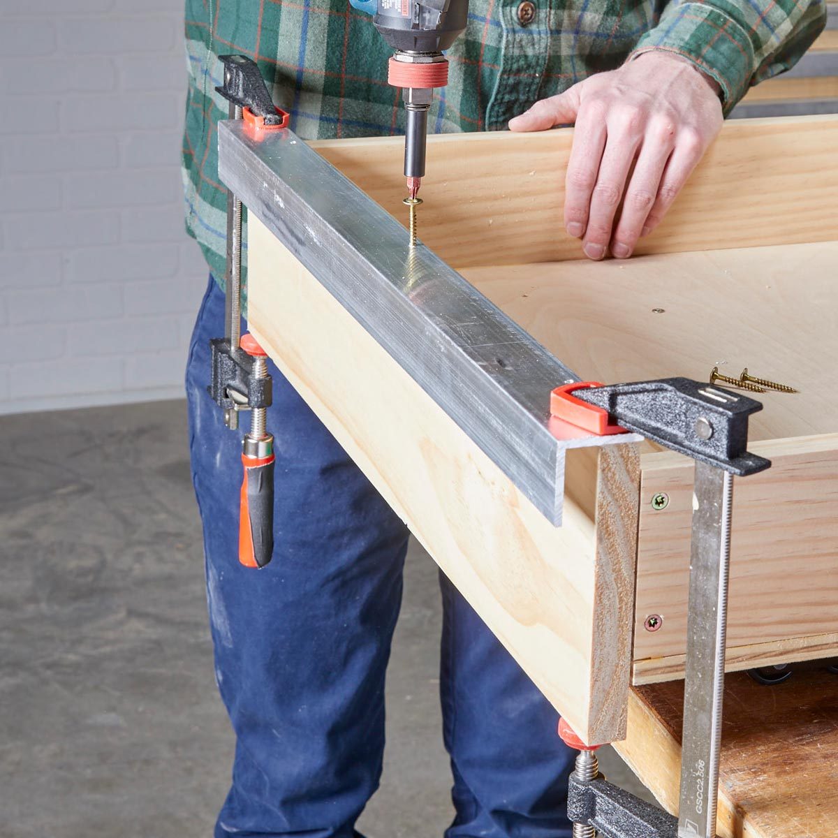 A person uses a power drill to secure screws into a wooden drawer, which is clamped to a work surface in a workshop.