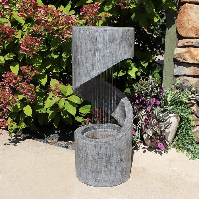 A modern, spiraled concrete fountain flows water into a circular basin, surrounded by vibrant green foliage and decorative plants in a sunny outdoor space.
