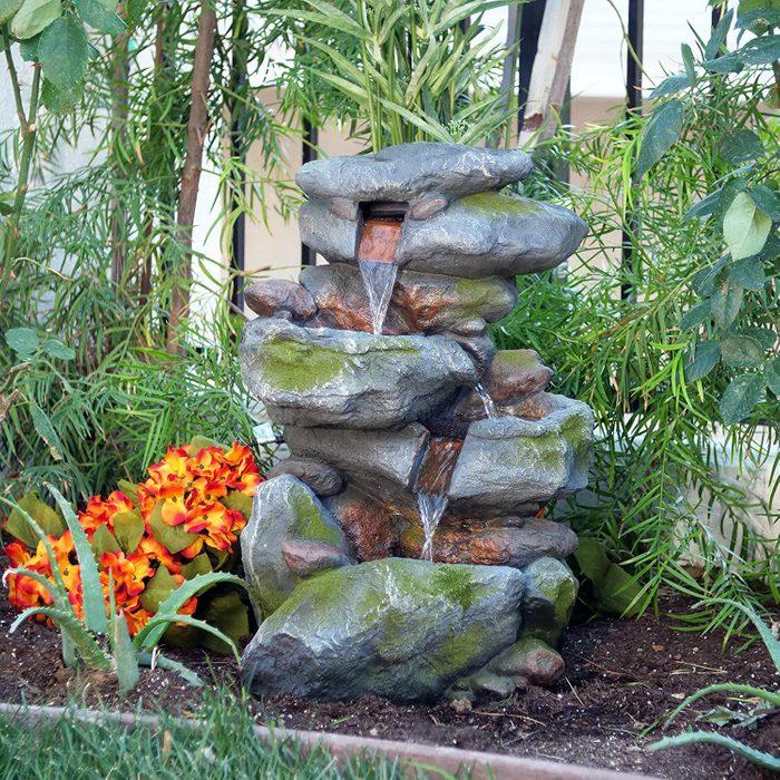 A stone water fountain cascades water down stacked rocks, surrounded by green plants and bright artificial flowers, enhancing the tranquil garden setting.
