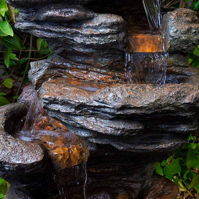 A stone fountain cascades water down multiple tiers, surrounded by lush green foliage, creating a natural, tranquil ambiance.