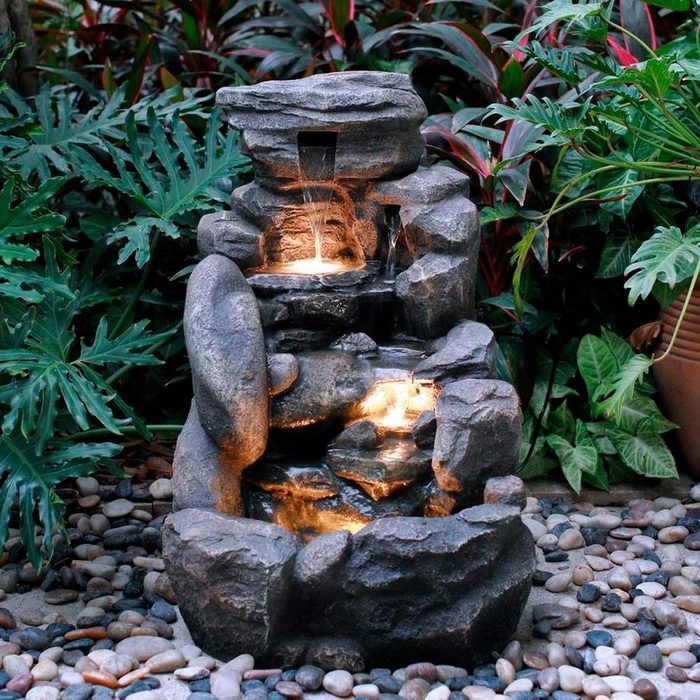 A decorative stone fountain cascades water between tiers, illuminated by lights, surrounded by lush green foliage and smooth pebbles.