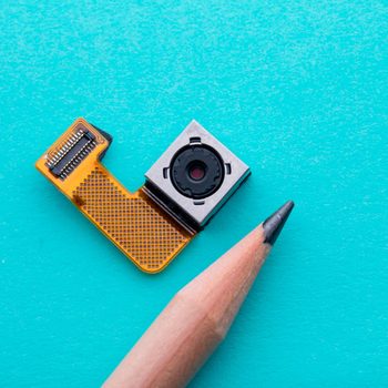 A small camera module with a yellow connector sits next to a sharpened pencil on a bright turquoise surface.