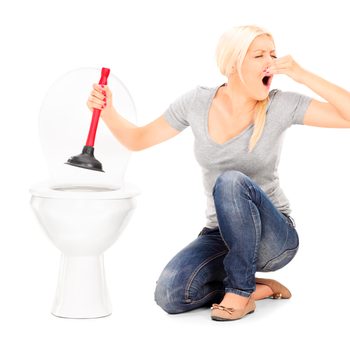 A woman kneels beside a toilet, holding a plunger while pinching her nose, expressing discomfort in a bright, uncluttered environment.