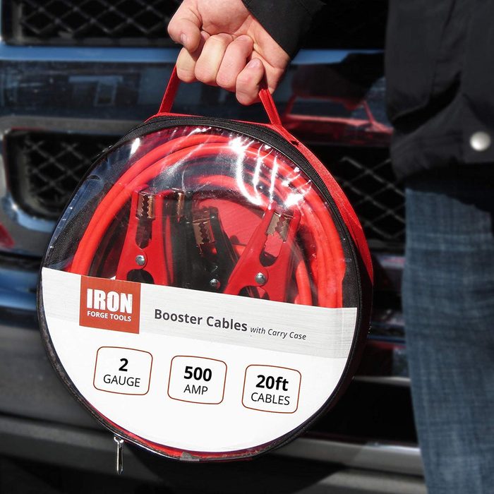 A person holds a circular case containing bright red booster cables, with visible metal clamps, in front of a vehicle's grille.