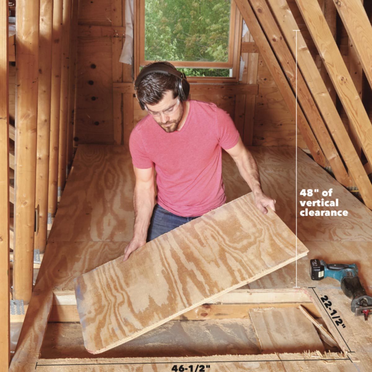 A man in a red shirt lifts a plywood piece inside a wooden attic, surrounded by beams and a bright window, with tools on the floor nearby.