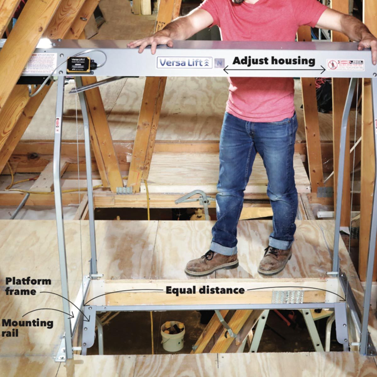 A person adjusts a lifting mechanism labeled "Versa Lift" in an attic, standing on a platform framed by wood beams and plywood flooring.
