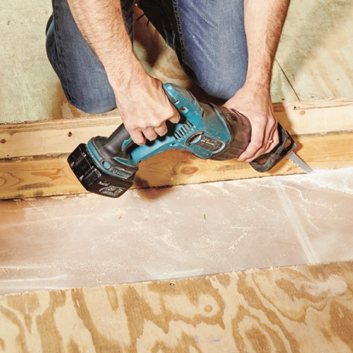 A person uses a power saw to cut through a wooden beam on a floor, surrounded by wood debris and a plywood base.