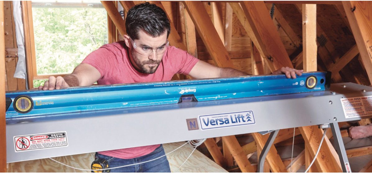 A man adjusts a level tool on a VersaLift in an attic, surrounded by wooden beams and natural light coming through a window.