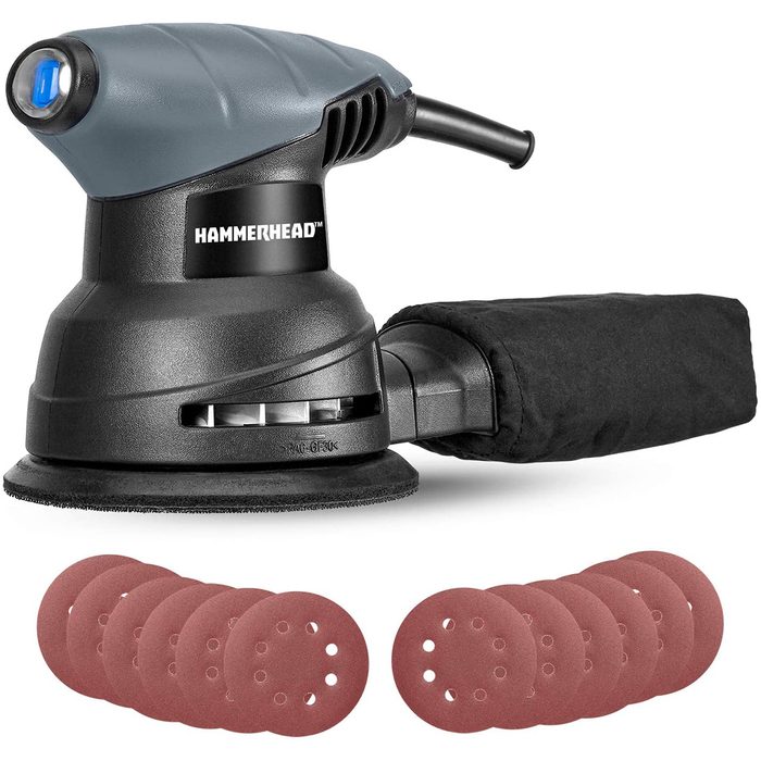 A black and gray power sander rests on a surface, ready for use. Red sanding discs are arranged in front, indicating preparation for a sanding task.