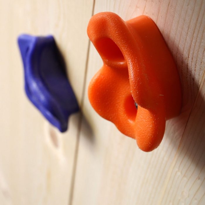 Two climbing holds, one orange and one purple, are mounted on a light wooden wall, suggesting a climbing environment.