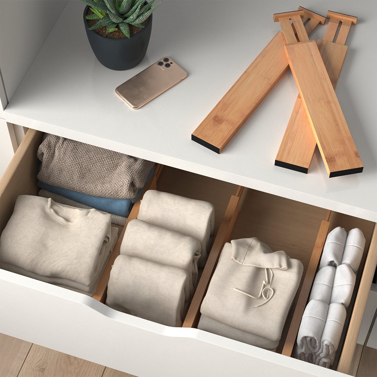 A neatly organized drawer holds folded clothes in compartments, while a potted plant and smartphone rest on a smooth surface nearby, with wooden planks beside them.