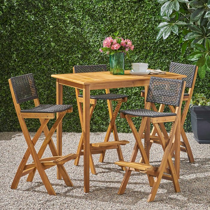 A wooden table with four folding chairs sits on gravel, surrounded by green foliage. A vase of pink flowers decorates the table alongside dishes.