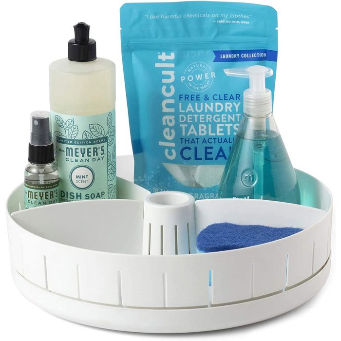 A white caddy holds cleaning products: mint dish soap, laundry detergent tablets, and a blue sponge, organized for efficient use in a cleaning context.