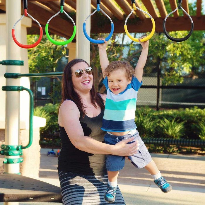 A smiling woman holds a joyful child swinging from hanging rings at a playground, surrounded by greenery and a sunny atmosphere.