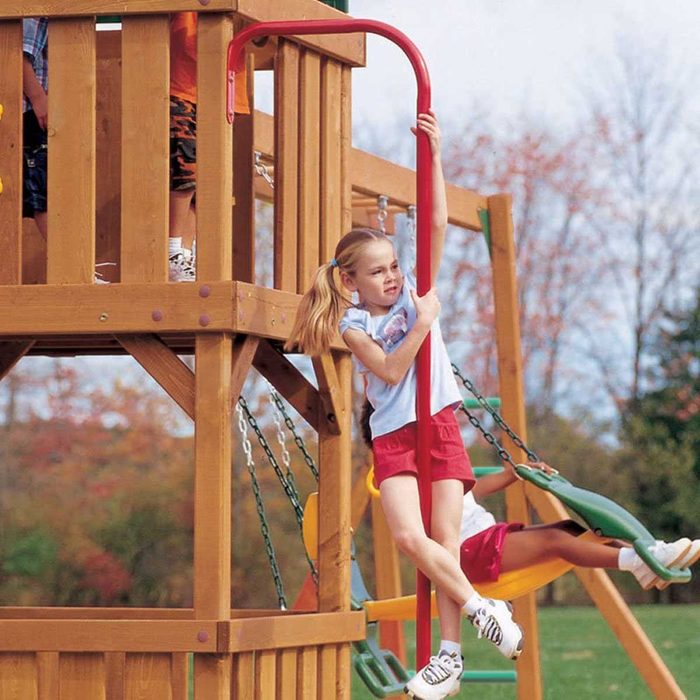 A girl hangs on a red pole, sliding down from a wooden playground structure while another child swings nearby in an outdoor park.