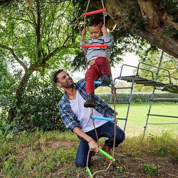 A man helps a child swing on a climbing rope, surrounded by grass and trees, with playground equipment visible in the background.